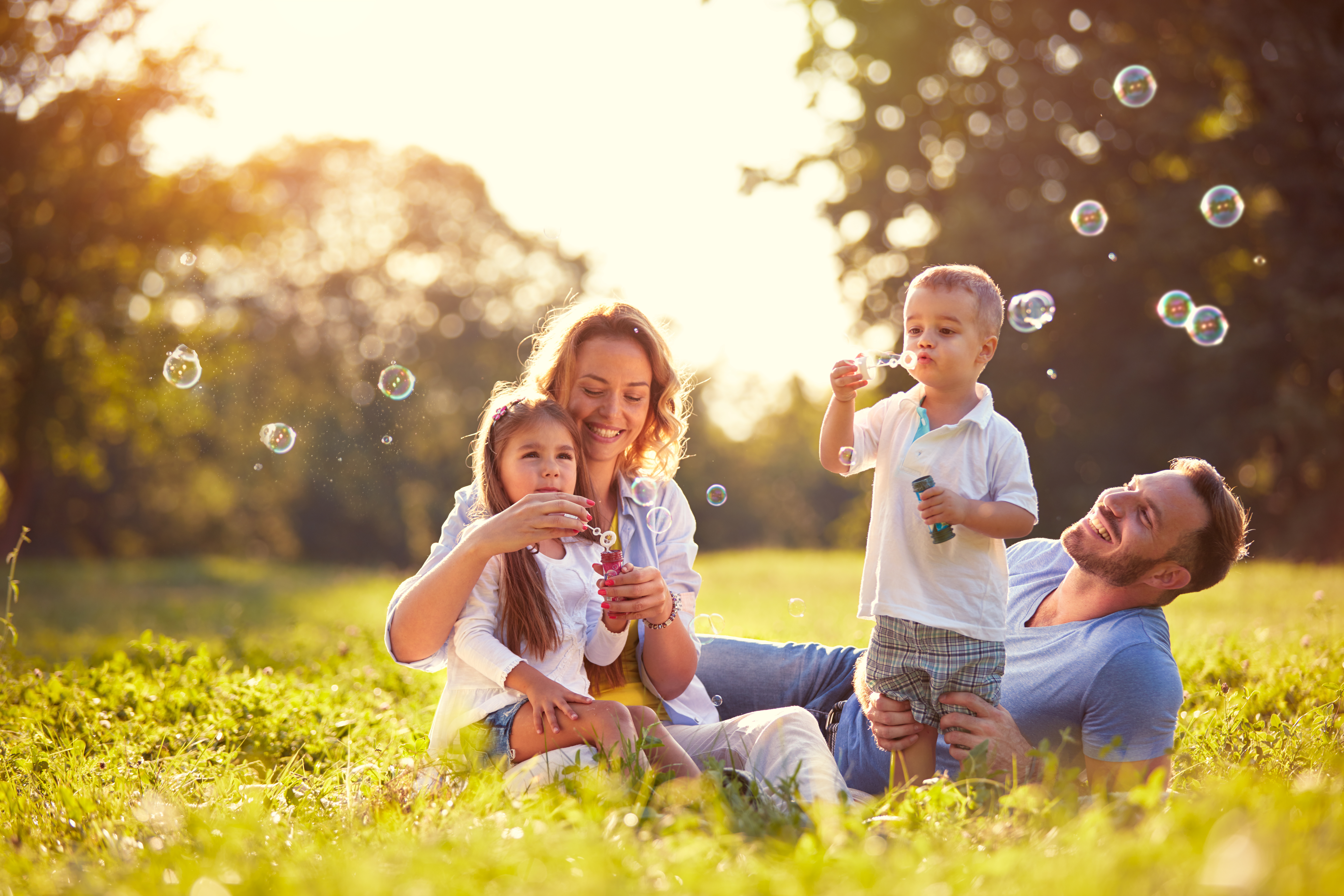 A family sits in a sunlit meadow blowing bubbles together