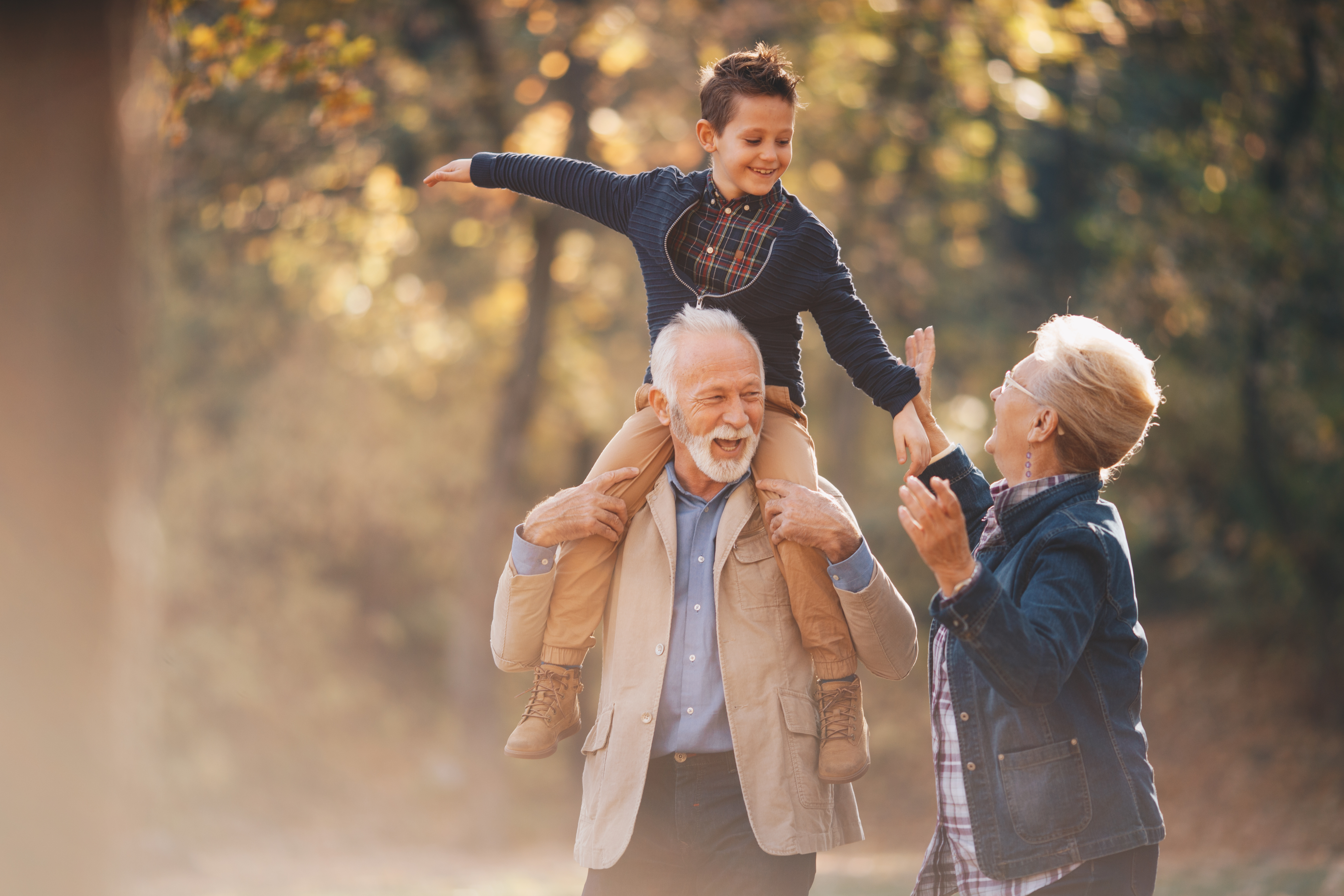 Grandparents laugh with a young boy sitting on his grandfather’s shoulders in autumn light