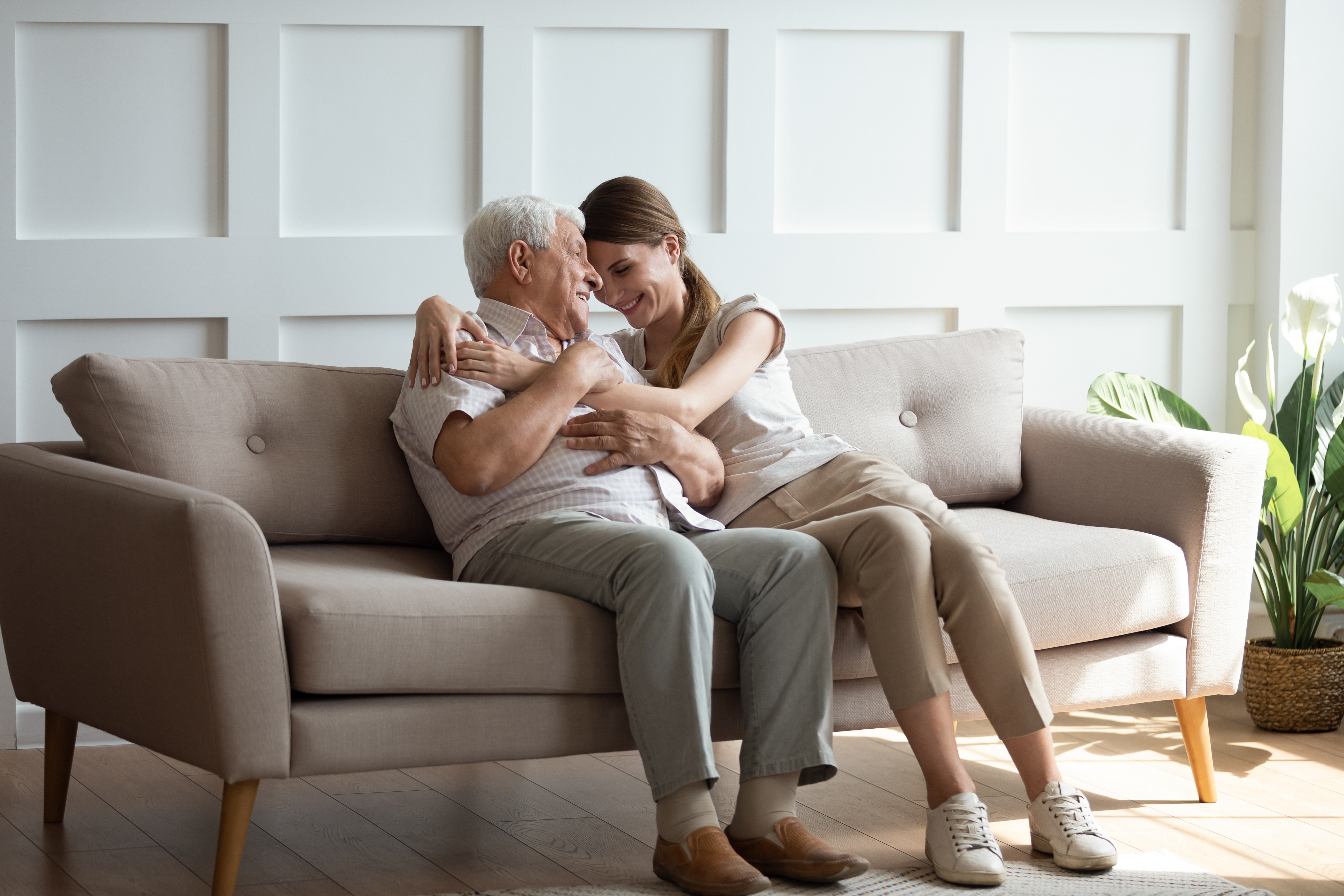 An older man and a younger woman sit together on a living-room sofa, hugging