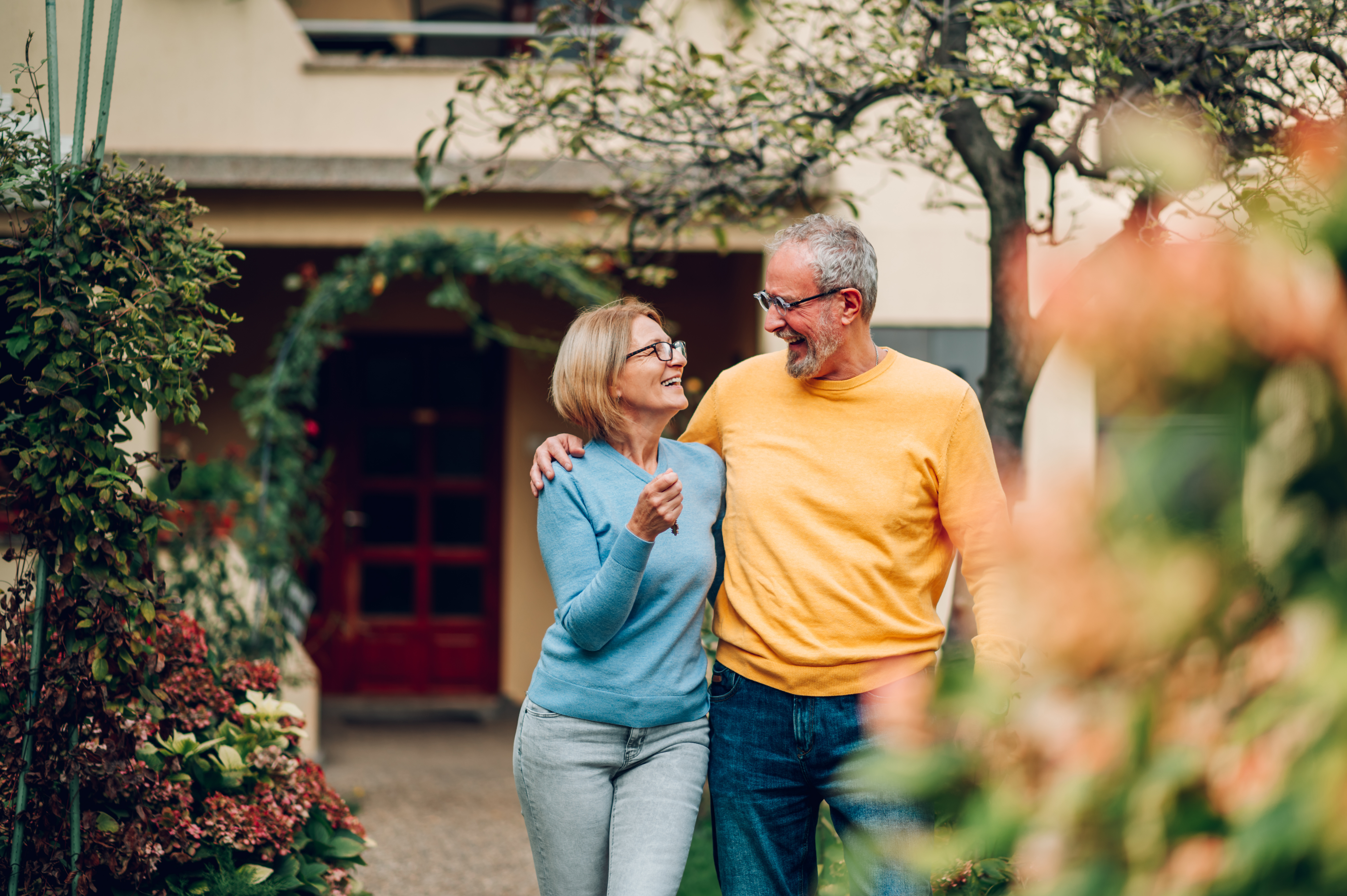 An older couple walk together outside a house, smiling, the woman holding a set of keys