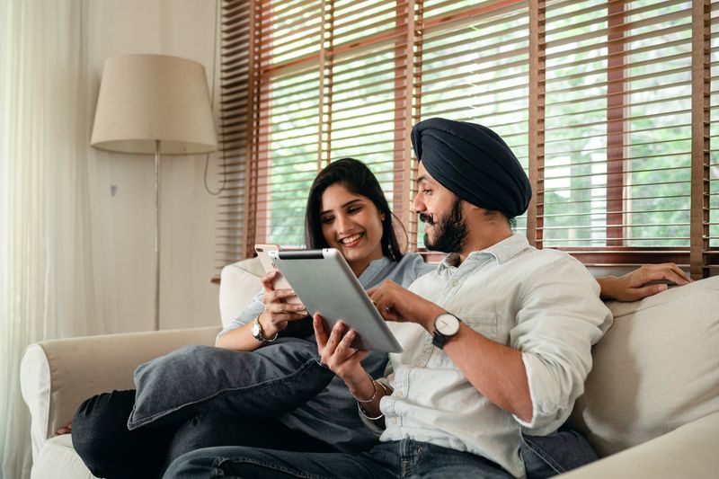 A couple sits on a sofa looking at a tablet together in a sunlit living room