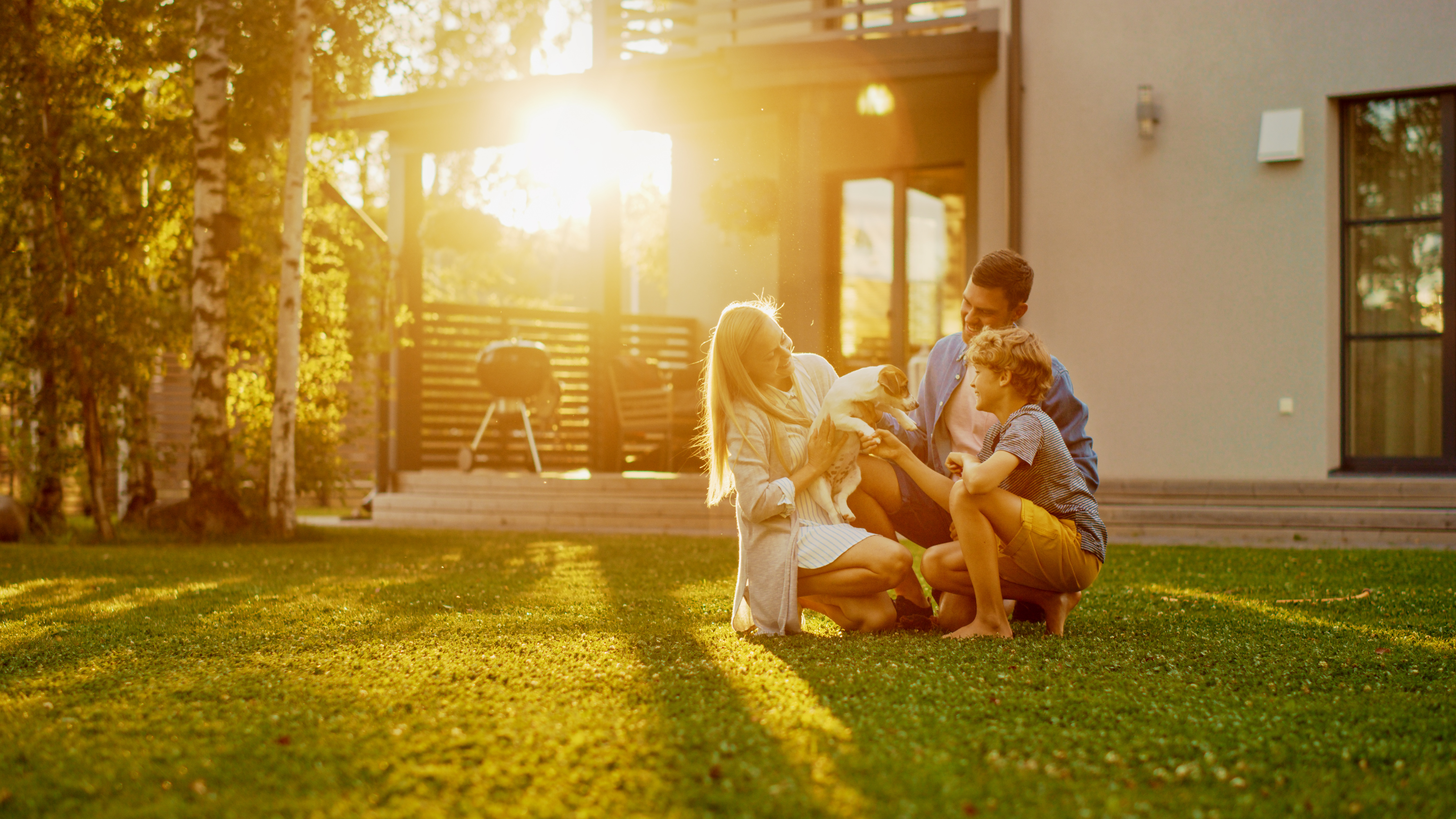 A mother, father, and young son kneel on the lawn with a puppy in front of their house