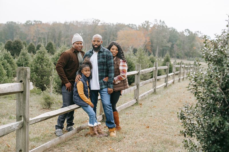 A family of four stands together at a tree farm, smiling in front of evergreen rows and a wooden fence